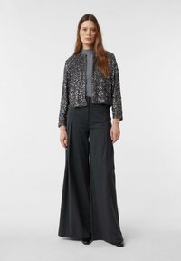 Femme portant une veste en sequins gris sur un haut gris, associée à un pantalon large taille haute gris foncé et des chaussures noires à bouts pointus.