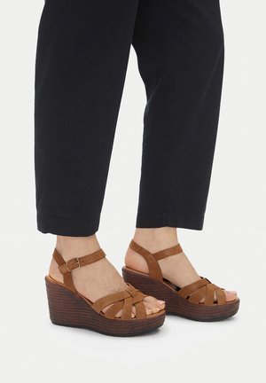 Pies con sandalias cuña de cuero marrón con tiras en el tobillo, combinadas con pantalones negros recortados sobre un fondo liso.