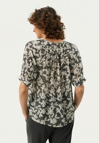 Gennemsigtig bluse med et sort blomsterdesign på en lys baggrund, korte optrallede ærmer og let rynket detalje ved nakken bagpå.