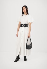 Vit lång klänning med puffärmar och knapprad fram. Svart nitarbälte som drar in midjan. Modellen håller en svart väska med silvernitar och bär spetsiga stövlar.