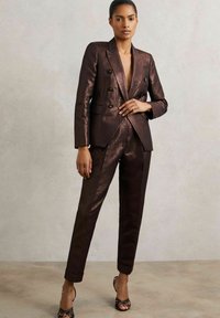 Tmavě bronzový oblek s přiléhavým dvouvrstvým sakem a na míru šitými kalhotami. Materiál má lesklý, texturovaný povrch. Kombinováno s černými sandály na podpatku se stužkami.