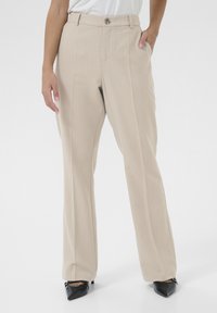 Beige gerade geschnittene Hose mit subtilen Streifen, ausgestattet mit einem vorderen Knopfverschluss und seitlichen Taschen, kombiniert mit schwarzen spitzen Absatzschuhen.