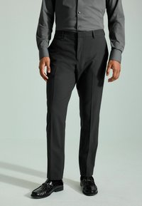 Pantalons de costume noirs à coupe ajustée, avec des plis sur le devant et des poches, associés à une chemise à boutons grise et des chaussures de ville noires.