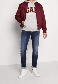 Uomo con giacca bomber bordeaux, felpa grigia con il testo "GAP New York", jeans blu arrotolati, sneaker bianche, in piedi con una mano in tasca.