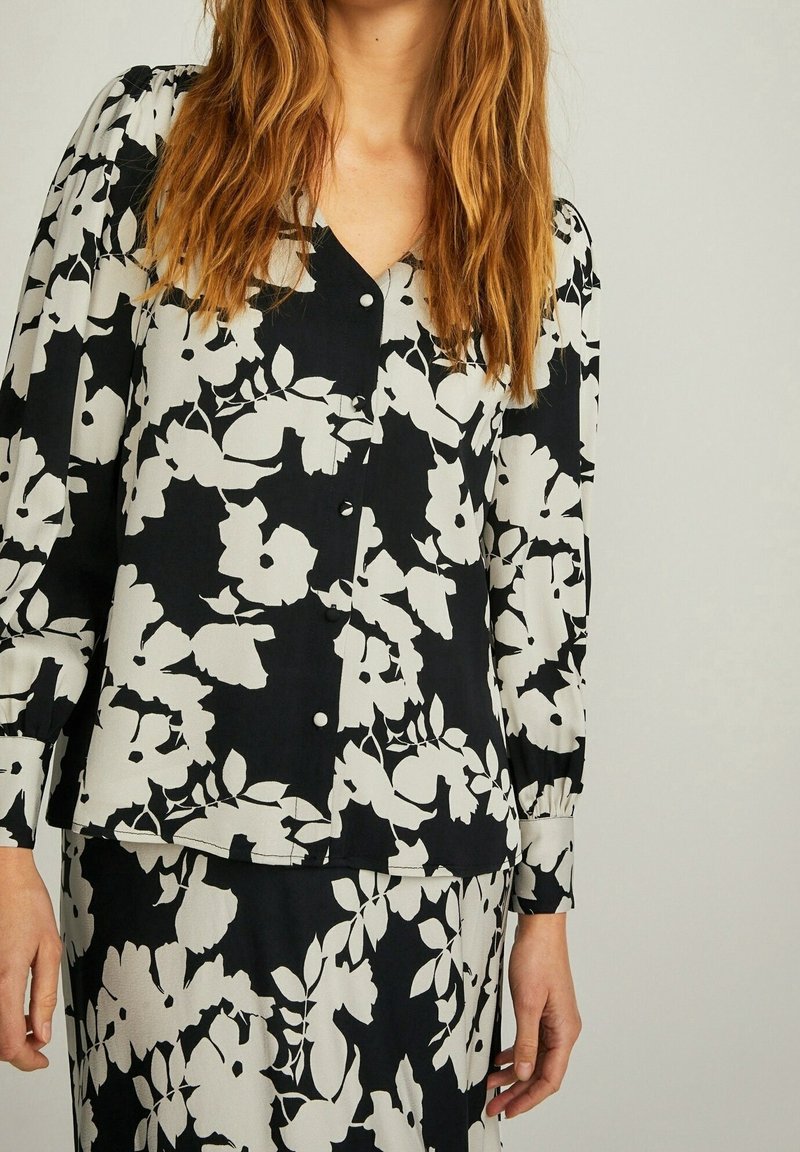 Femme portant un chemisier boutonné à motif floral noir et blanc et une jupe assortie, avec de longs cheveux roux détachés, debout devant un fond uni.