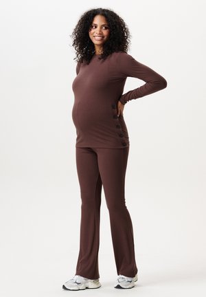 Zwangere vrouw in een bruin topje met lange mouwen en broek, staand met hand op de rug en glimlachend, draagt witte sneakers op een witte achtergrond.