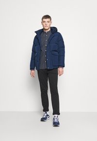 Marine blauwe pufferjack met een capuchon, voorzien van een rits en drukknoopsluiting, gecombineerd met een gepunte blouse en zwarte broek. Zwart en blauwe sneakers.