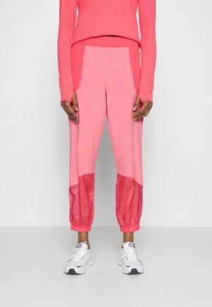 Person wearing pink jogger pants with darker pink mesh panels at the knees and white sneakers, standing against a plain white background.