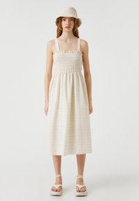 Vaalean beige mekko, jossa on kootut rintakappaleet, henkselit ja kerrottu hame. Yhdistä se sopivan ämpärilakin ja korkokenkien kanssa.