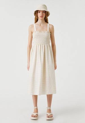 Vaalean beige mekko, jossa on kootut rintakappaleet, henkselit ja kerrottu hame. Yhdistä se sopivan ämpärilakin ja korkokenkien kanssa.