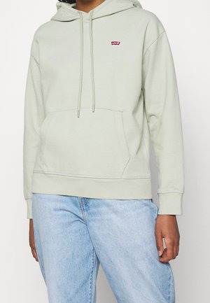 Personne portant un sweat à capuche Levi's gris clair avec poche avant et cordons, assorti à un jean bleu clair, sur fond uni.