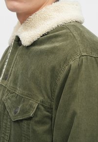 Gros plan d'une veste en velours côtelé verte avec un col sherpa blanc et des boutons-pression en métal sur la poche poitrine et à l'avant.