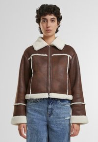 Giacca cropped in pelle marrone con un morbido colletto e profili in shearling crema, dotata di chiusura con zip e grandi tasche frontali.