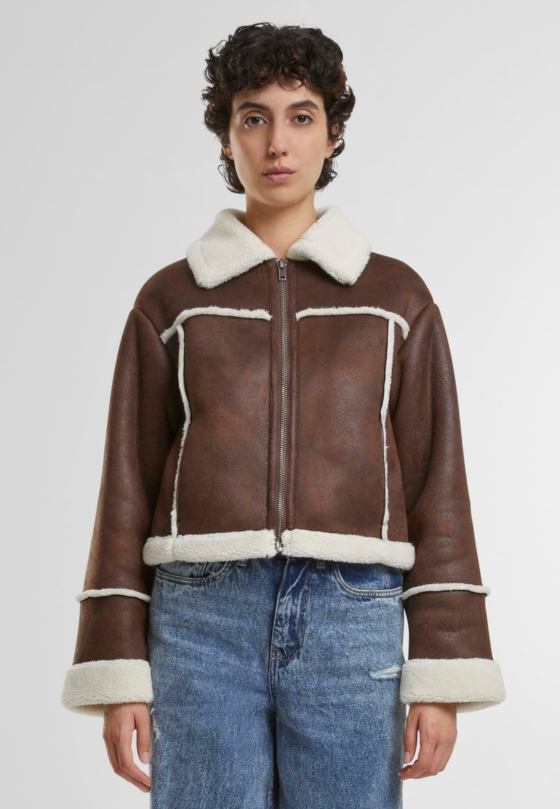Giacca cropped in pelle marrone con un morbido colletto e profili in shearling crema, dotata di chiusura con zip e grandi tasche frontali.
