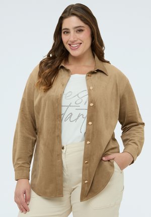 Donna sorridente con lunghi capelli castani che indossa una camicia color sabbia abbottonata sopra una maglietta bianca con stampa e pantaloni beige chiaro, con una mano in tasca.