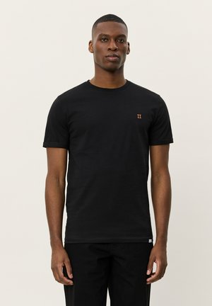 Homme debout de face, portant un t-shirt noir uni avec un petit symbole orange sur la poitrine et un pantalon noir, fond neutre.
