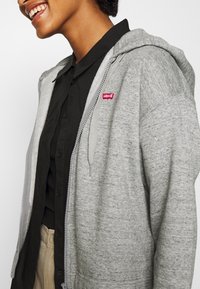 Personne souriante portant une chemise noire boutonnée, un sweat à capuche Levi's gris clair zippé et un pantalon beige, montrée de la nuque jusqu'en bas.