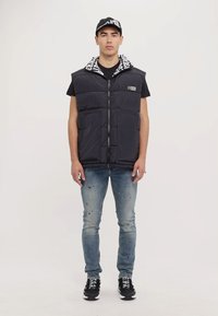 Gilet rembourré noir avec fermeture éclair à l'avant, comportant une doublure de capuche à motifs, porté sur une chemise noire, associé à un jean bleu déchiré et des baskets noires.