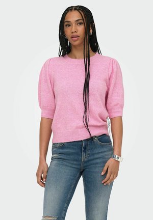 Femme aux longs cheveux tressés portant un pull rose à manches courtes et un jean bleu, debout devant un fond uni.