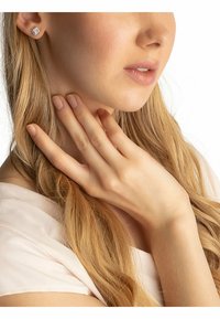Orecchini a bottone rotondi di tono argentato con piccoli cristalli trasparenti incastonati in una superficie testurizzata. La mano della modella poggia vicino al collo.