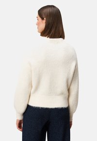 Kremowy, puszysty sweter z ribbowanym dołem i mankietami, o wysokim kołnierzu i luźnym kroju. Materiał wydaje się miękki i teksturowany.