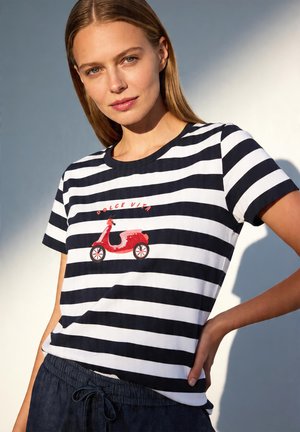 Kobieta z prostymi blond włosami, ubrana w czarno-białą koszulkę w paski z czerwonym skuterem i napisem "Dolce Vita" z przodu.