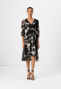 Robe noire portefeuille avec un motif floral dans des nuances de beige, blanc et marron. Manches bouffantes courtes et ourlet asymétrique. Portée avec des talons noirs à lanières.