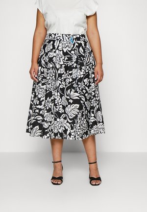 Femme portant un haut blanc avec une jupe midi à fleurs noire et blanche et des talons ouverts noirs avec bride à la cheville, debout devant un fond uni.