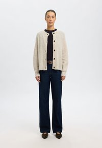 Beige, fuzzy cardigan met ronde halslijn, knopen aan de voorkant en geribbelde mouwen, gecombineerd met donkerblauwe high-waisted jeans en bruine schoenen.