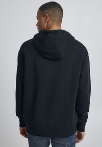 Sudadera negra con capucha, puños y dobladillo acanalados, con un ajuste holgado y una textura suave. Se muestra desde la parte posterior de una persona que lleva jeans.