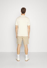 Béžová polo košile a svetle khaki kraťasy. Košile má límec a krátké rukávy. Celý outfit doplňují bílé tenisky a kotníkové ponožky.