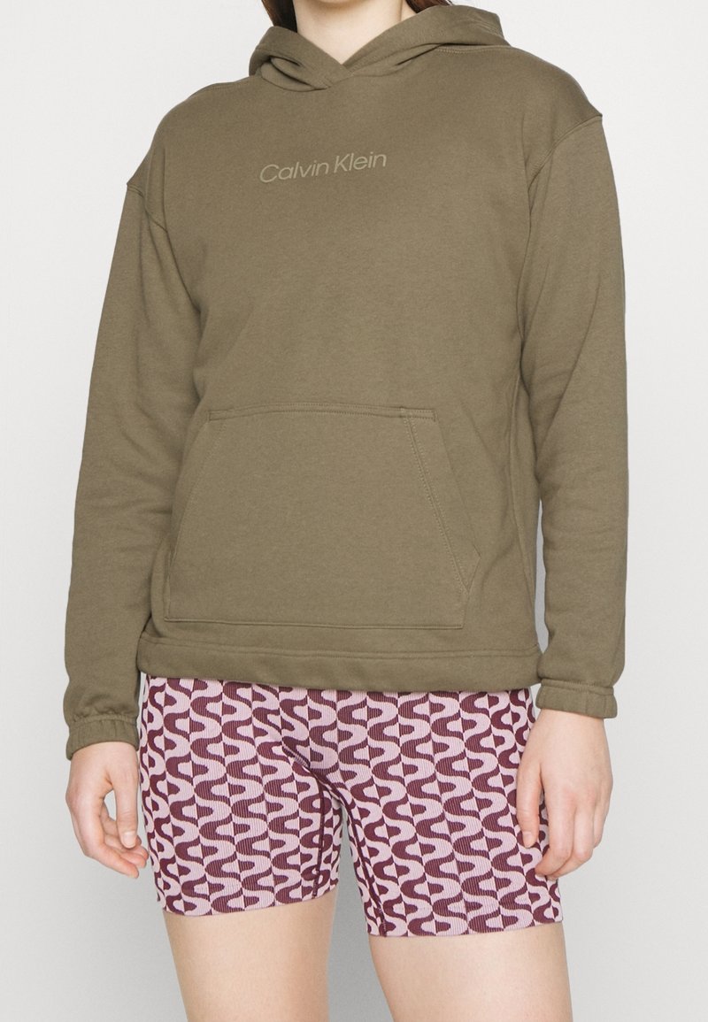 Personne portant un sweat à capuche Calvin Klein vert olive et un short rose à motifs géométriques, debout devant un fond uni.