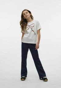 Vit bomullströja med ett röd och svart bandtryck på framsidan, tillsammans med mörkblå vida byxor och svarta sneakers.