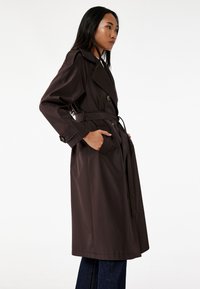 Trench coat marrón oscuro con cintura ceñida por cinturón, grandes botones y bolsillos laterales. Presenta un clásico cuello de solapa y una textura suave.