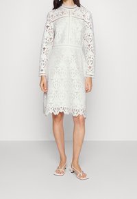 Bílé krajkové šaty s dlouhými rukávy a vlnkovým lemem, zdobené květinovými vzory a přiléhavým střihem. Spárovány s bílými sandály na páscích.