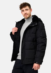 Schwarze Pufferjacke mit einem fellgefütterten Kapuzenkragen, ausgestattet mit einem Reißverschluss, Druckknöpfen und strukturierten Schulterakzenten. Getragen über einem grauen Rollkragenpullover.