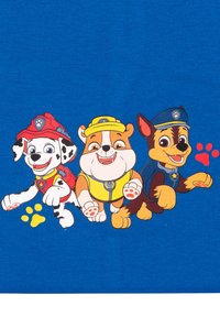 Tre cani animati in uniformi da soccorso—pompieri, costruzione e polizia—stanno correndo con impronte di zampe colorate dietro di loro su uno sfondo blu.
