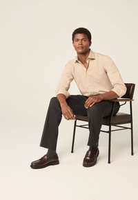 Béžová košile na knoflíky, tmavě šedé kalhoty na míru, hnědé loafers, sedící na černé kovové židli s dřevěným opěrátkem. Neutrální pozadí.