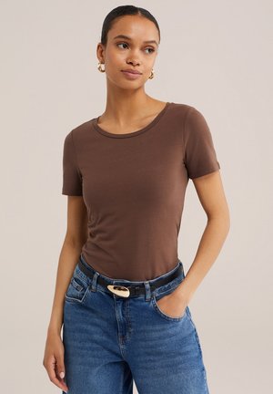 Kvinde iført simpel brun kortærmet top, blå jeans med sort bælte og guldringe i ørerne, stående med den ene hånd i lommen.