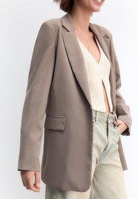Blazer su misura marrone chiaro con rever a punta, chiusura a singolo bottone, tasche laterali e una texture di tessuto liscia, abbinato a un top lavorato a maglia.