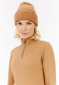 Bruine gebreide beanie met een geribd patroon, gecombineerd met een lichtbruine longsleeve top met een ritssluiting bij de hals en subtiel logo detail.