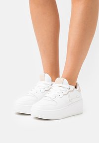 Białe trampki ze gładkim wykończeniem ze skóry, wyściełanym językiem i teksturowanymi akcentami w kolorze beżowym. Posiadają grubą podeszwę na platformie oraz perforowane detale.