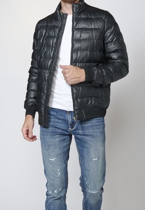 Mand iført sort quiltet jakke over hvid skjorte med blå slidte jeans, justerer jakke lynlås.