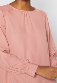 Blusa rosa chiaro con scollo rotondo, scollatura arricciata e maniche lunghe con polsini abbottonati e sottili pieghe lungo le maniche.