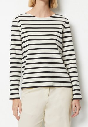Vrouw draagt een zwart-wit horizontaal gestreept shirt met lange mouwen en lichtbeige broek, staand tegen een witte achtergrond.