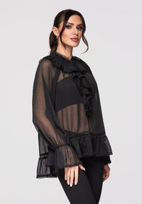 Femme portant un chemisier noir transparent avec col et poignets à volants, assorti d'un pantalon noir, debout devant un fond clair.