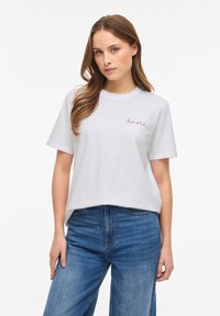 Νεαρή γυναίκα με μακριά καστανά μαλλιά που φοράει λευκό t-shirt με το κείμενο "amore" και μπλε τζιν, στέκεται μπροστά σε απλό λευκό φόντο.