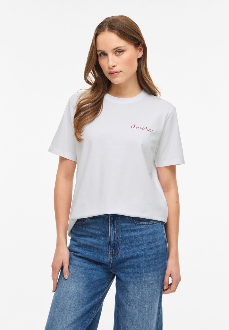 Νεαρή γυναίκα με μακριά καστανά μαλλιά που φοράει λευκό t-shirt με το κείμενο "amore" και μπλε τζιν, στέκεται μπροστά σε απλό λευκό φόντο.