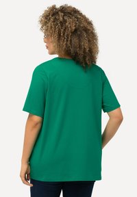 Camiseta verde de manga corta con cuello redondo, que presenta una textura suave y un detalle de costura en la parte trasera. Se lleva con pantalones oscuros.