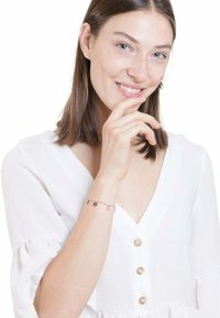 Pulsera de oro y negro en una muñeca, con encantos circulares. El modelo lleva una blusa blanca con mangas abullonadas y botones.